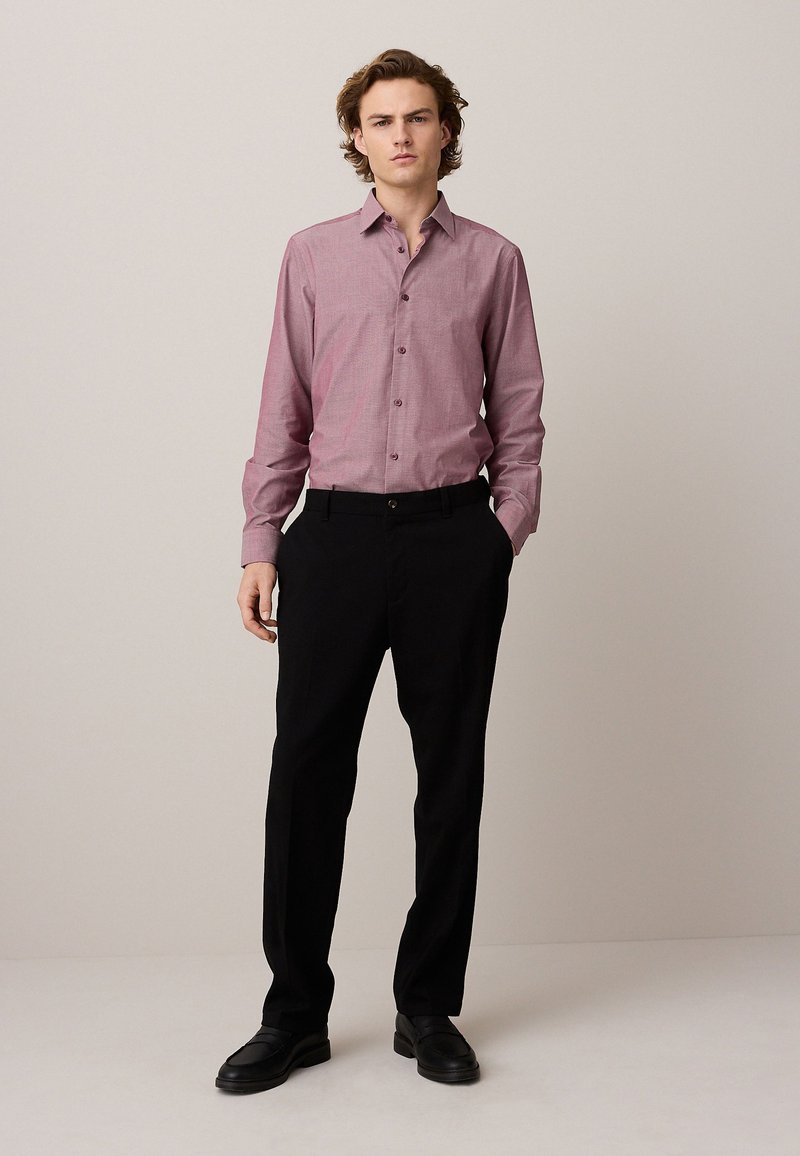 Giovane uomo in piedi con le mani in tasca che indossa una camicia bordeaux con bottoni, pantaloni neri e scarpe nere su uno sfondo semplice.