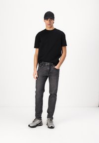 Svart t-skjorte, mørkegrå jeans og sølvfargede treningssko. Modellen har på seg en svart caps og står mot en hvit bakgrunn. Enkel design.