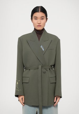 Blazer verde oliva con un design strutturato, due tasche anteriori e una cintura in vita. Presenta ricami floreali sulla manica sinistra.