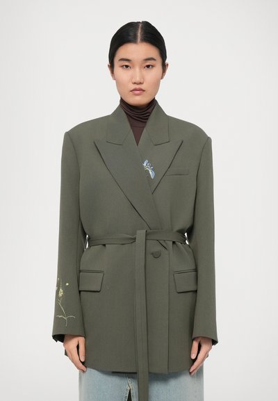 Olivengrøn blazer med en struktureret design, to forlommer og et taljebånd. Har blomsterbroderi på venstre ærme.