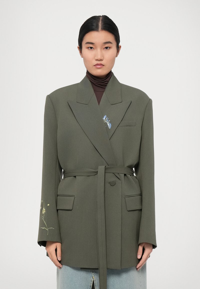 Blazer verde oliva con un design strutturato, due tasche anteriori e una cintura in vita. Presenta ricami floreali sulla manica sinistra.