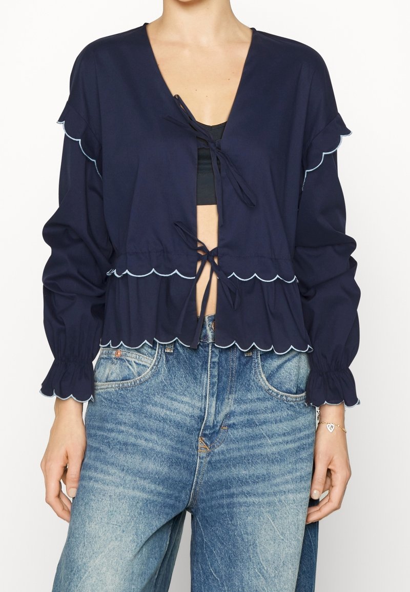 Femme portant un chemisier court bleu marine avec des bords festonnés et des fermetures à nouer, associé à un jean bleu clair taille haute.