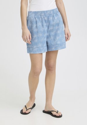 Partie inférieure d'une personne portant un short bleu clair brodé de fleurs, un haut blanc et des tongs noires sur un fond uni.