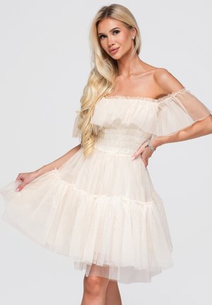 Blond kvinde poserer iført off-shoulder, cremefarvet, flæset tylskjole med langt bølget hår og et armbånd på håndleddet.