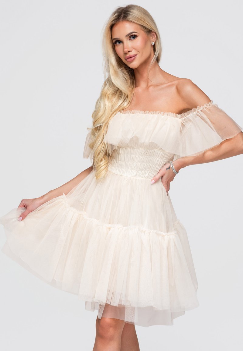Femme blonde posant avec une robe en tulle crème à volants et épaules dénudées, cheveux longs ondulés, et un bracelet au poignet.