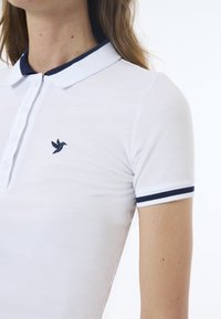 Polo bianco con colletto e polsini blu navy, caratterizzato da un logo di uccello blu navy sul lato sinistro del petto. Realizzato in tessuto testurizzato, design a maniche corte.