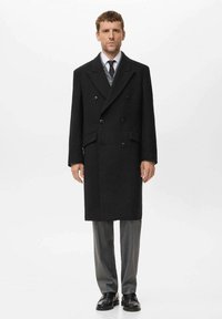 Doppelt breiter schwarzer Wollmantel mit schlanker, taillierter Passform, Reverskragen und seitlichen Taschen, kombiniert mit grauen Hosen und schwarzen Schuhen.