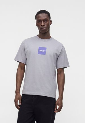 Muškarac koji nosi svijetlosivu majicu s ljubičastim kvadratnim logotipom i bijelim tekstom "huf", stoji na bijeloj pozadini.