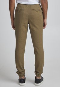 Pantalones jogger color caqui con cintura elástica, tobillos ajustados y dos bolsillos traseros con detalles de botón y cremallera. Textura suave.