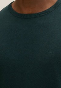 Maglione lavorato a maglia di un verde scuro con scollo rotondo. Presenta un logo discreto sul davanti, realizzato in un tessuto morbido e strutturato.