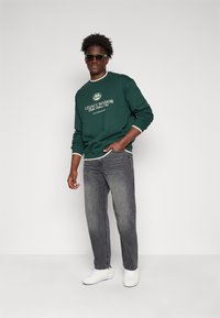 Felpa verde con dettagli bianchi e logo "LEGACY MANOR", abbinata a jeans grigi e sneakers bianche. Il modello indossa occhiali da sole.