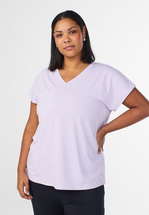 Mustade sirgete juustega naine kannab helelilla V-kaelusega T-särki ja musti pükse, üks käsi puusal, taust on ühevärviline.