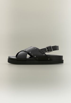 Schwarze Ledersandale mit gekreuzten Riemen, kleinen Metallnieten und verstellbarer Schnalle auf flacher Sohle vor hellem Hintergrund.