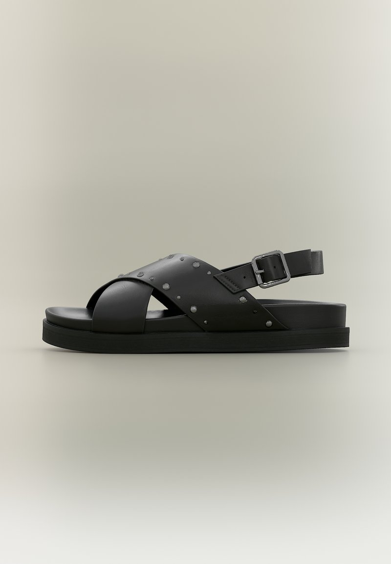 Μαύρο δερμάτινο σανδάλι με σταυρωτούς ιμάντες, μικρές μεταλλικές τρουκς και ρυθμιζόμενο πόρπη σε επίπεδη σόλα σε ανοιχτό φόντο.