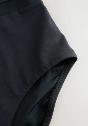 Artimiausias juodų audinių maudymosi trumpikių vaizdas su matomomis siūlėmis ir juosmens diržu ant šviesaus fono.