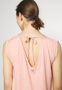 Blusa rosa dal taglio morbido, con maniche corte a balze, scollo a V sul retro con chiusura a lacci e motivi floreali bianchi su tutta la superficie.
