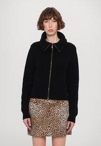 Svart ribbat stickat jacka med en stor krage och guld blixtlås, kombinerat med en leopardmönstrad minikjol med svarta fläckar på en beige bakgrund.