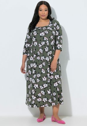 Frau mit langen dunklen Haaren trägt ein grünes Midikleid mit weißem Blumenmuster und rosa flache Schuhe, steht vor einer weißen Wand.