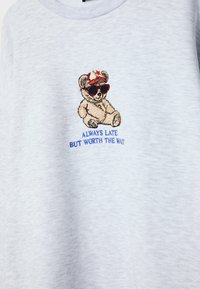 Chemise gris clair avec un ours en peluche brodé portant une casquette rouge et des lunettes de soleil au-dessus du texte « Always late but worth the wait » en bleu.