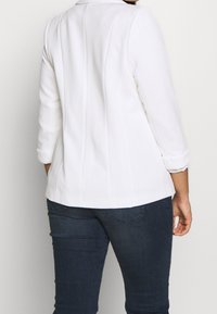 Bílá žebrovaná bunda s obtaženým střihem, tříčtvrtečními rukávy a kulatým límcem, zkombinovaná s tmavě modrými upnutými džínami.