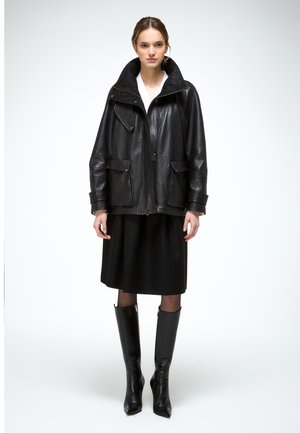 Femeie purtând o geacă din piele neagră supradimensionată, fustă midi neagră, ciorapi transparenți și cizme negre înalte până la genunchi, cu vârful ascuțit, stând pe un fundal alb.