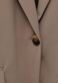 Blazer marron avec un bouton de couture rond brun foncé fixé près du revers et de la zone du col.