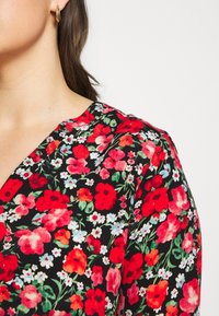 Femme portant une robe noire avec un motif floral rouge, rose et blanc, et une petite boucle d'oreille en or en forme d'anneau, montrée du cou à l'épaule sur fond blanc.