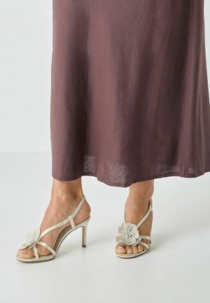 Fødder iført beige højhælede sandaler med blomstermotiver, delvist dækket af en brun nederdel, der går til anklerne.