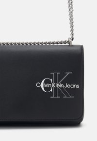Czarna skórzana torba na ramię z srebrnym łańcuszkiem. Posiada wytłoczone białe logo "Calvin Klein Jeans". Gładka tekstura i prostokątny kształt.