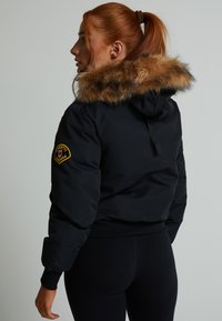 Félbevágott fekete dzseki műszőrme kapucnival, elasztikus mandzsettákkal és aljjal, valamint egy feliratos folttal az ujján. Sima textúra és testhezálló kialakítás.