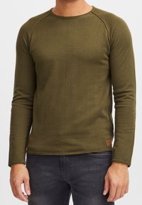 Chemise en maille à manches longues vert olive avec une texture côtelée, un col rond et des manches raglan. Elle présente un petit écusson logo marron à l'ourlet.