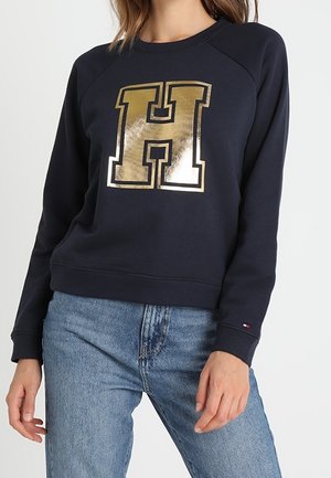 Marineblå sweatshirt med et guldmetalisk "H" grafik på fronten, med raglanærmer og ribbede manchetter, kombineret med lyseblå jeans.