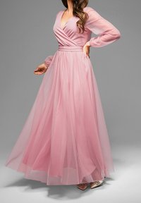 Femme portant une longue robe en tulle rose avec un corsage ajusté, un décolleté en V et des manches longues transparentes sur un fond gris.