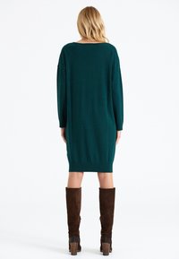 Robe-pull tricotée couleur sarcelle avec une coupe ample, manches longues, et ourlet côtelé. Associée à des bottes marron molletonnées montantes jusqu'aux genoux.