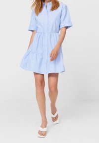 Abito camicia in cotone azzurro chiaro con maniche corte, chiusura a bottoni nella parte anteriore, vita elastica, gonna a strati e vestibilità comoda. Indossato con sandali bianchi.