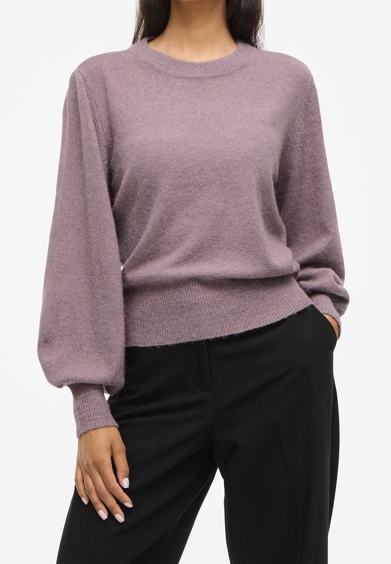Femme portant un pull mauve à manches longues avec poignets et ourlet côtelés, associé à un pantalon noir, debout avec une main dans la poche.
