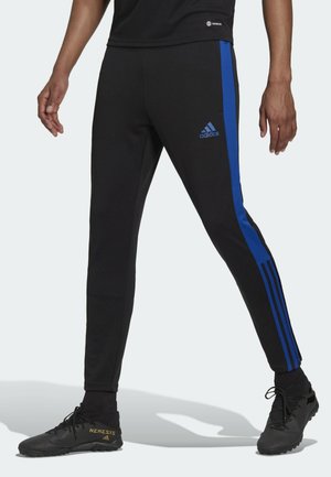 Muž nosící černé tepláky Adidas s modrými pruhy a černé fotbalové boty Adidas stojí s roztaženýma nohama.