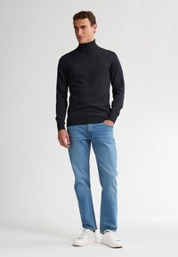 Czarny sweter z golfem o dopasowanym kroju, sparowany z jasnoniebieskimi dżinsami i białymi sneakersami, stojący na jednolitym tle.