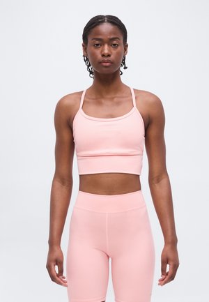 Femme aux cheveux tressés portant un crop top de sport rose clair et un short cycliste taille haute assorti, debout devant un fond uni.