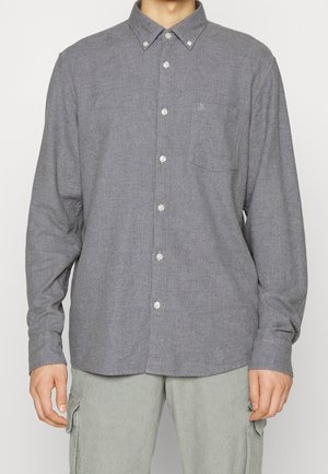 Uomo che indossa una camicia grigia a maniche lunghe con taschino sul petto e pantaloni cargo grigio chiaro su uno sfondo semplice.