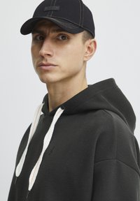 Felpa nera con cappuccio e lacci bianchi, con un logo ricamato. Indossata con un berretto nero con visiera cucita e un'etichetta sulla parte anteriore.