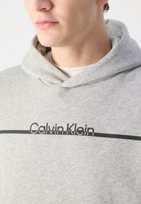 Grå hettegenser med snøring og en svart horisontalstripe med "Calvin Klein"-tekst. mykt stoff med en glatt tekstur.