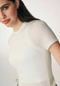 Helles beiges, geripptes T-Shirt mit kurzen Ärmeln, runder Halslinie und weicher Textur, das über subtile Nähte und eine schmale Passform verfügt.