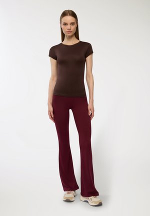 Top aderente marrone a maniche corte abbinato a pantaloni svasati bordeaux e sneakers in tonalità neutra, su uno sfondo semplice.