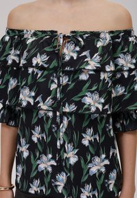 Blomstret bluse med avskåret skulder og lagdelte volanger. Svart stoff med hvite og blå blomster mønstre og grønne blader. Justerbar knyte detalj.