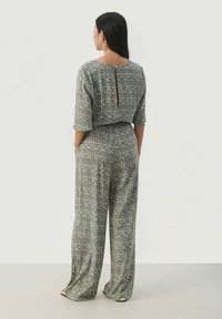 Zelena vzorčasta kombinezona s širokimi hlačami in odprtjem na hrbtu. Tkanina ima gladko teksturo z abstraktnim dizajnom.