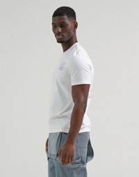Uomo che indossa una maglietta bianca con grafica e pantaloni blu a righe, in piedi di lato contro uno sfondo leggero e uniforme.