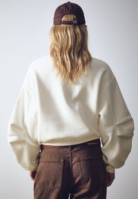 Felpa oversize color crema con maniche a sbuffo, abbinata a pantaloni marroni a vita alta. Completa il look un berretto marrone indossato al contrario.