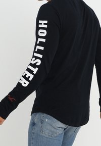 Black long-sleeve shirt featuring white "HOLLISTER" text along the left sleeve and a small red logo on the right cuff. Cotton material.