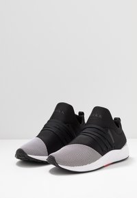 Schwarze Sportschuhe mit elastischen Riemen und einem strukturierten grauen Netz-Zehenbereich. Sie verfügen über eine glatte Silhouette und eine kontrastierende weiße Sohle.